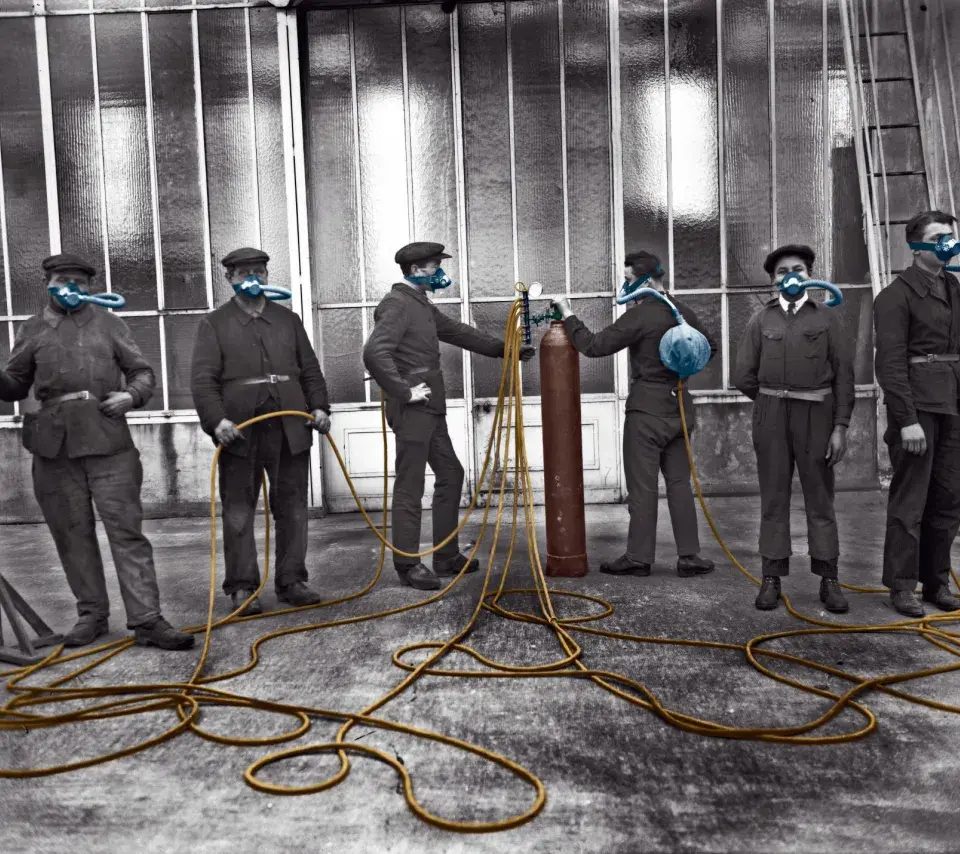 photo en noir et blanc colorisée. Plusieurs soldats avec chacun un masques à gaz relié à une bouteille collective (juin 1927). Destiné aux armées, ce dispositif devait prémunir les troupes des gaz de combat. Office national des recherches scientifiques et industrielles et des inventions.