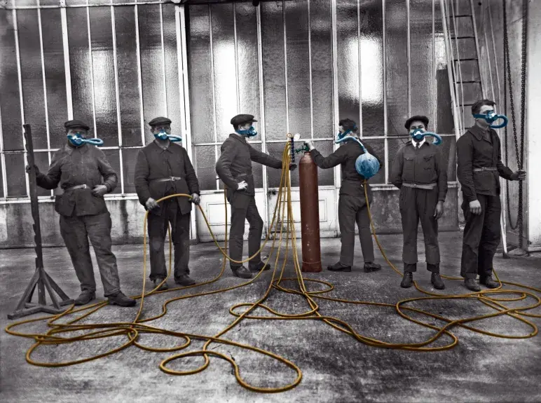 photo en noir et blanc colorisée. Plusieurs soldats avec chacun un masques à gaz relié à une bouteille collective (juin 1927). Destiné aux armées, ce dispositif devait prémunir les troupes des gaz de combat. Office national des recherches scientifiques et industrielles et des inventions.