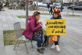 Deux femmes parlant assises sur des chaises sur une place, l'une d'elle, avec couronne de fleurs, tenant une pancarte "Raconte-moi une histoire d'animal"