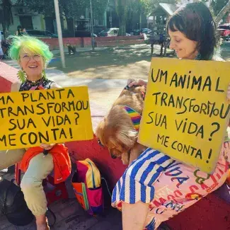 Deux femmes assises sur une place, tenant chacune une pancarte, "Uma planta transformou sua vida ? Me conta !" et "Um animal transformou sua vida ? Me conta !"