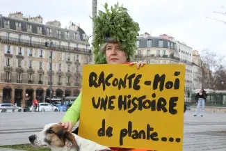 Femme sur une place, avec des feuilles dans les cheveux, caressant un chien, tenant une pancarte "Rcaonte-moi une histoire de plante"