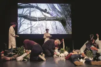 image de la performance "Arboretum-performance pour humains et plantes, Théâtre de la Resserre, CIUP, Paris, 25/10/18, (c) Hantu. Sur un scène, deux femmes en blouses debout, lisent. Quatre personnes agenouillées qui mettent des arbustes sur des personnes allongées avec des bandages. 